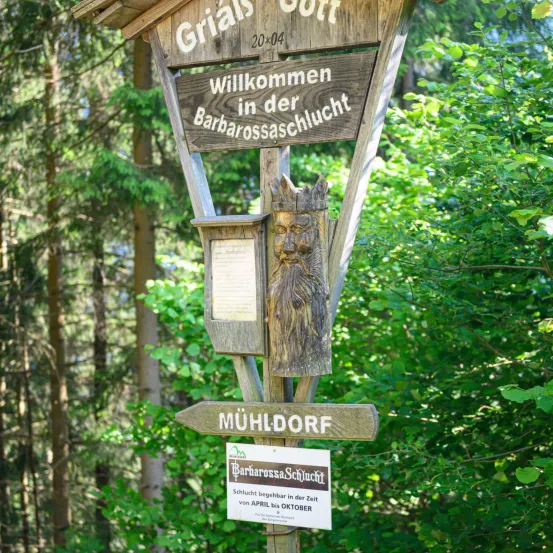 Ein hölzernes Schild heißt Besucher in der Barbarossa-Höhle von April bis Oktober willkommen. Es zeigt eine geschnitzte Figur und einen Wegweiser nach Mühldorf.