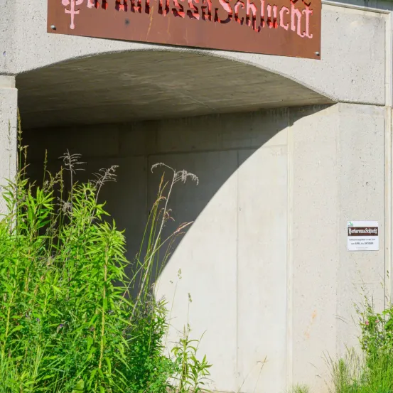 Ein Betonbogen mit einem Schild, das "Paradiso-Schlucht" darüber steht. Unter dem Bogen sind üppiges Grün und Pflanzen zu sehen.