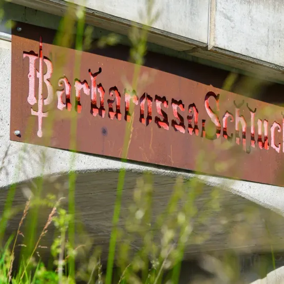 Ein Schild mit der Aufschrift 'Bartholomeus-Scharrer' ist an einem Gebäude montiert. Das Schild besteht aus rostigen Metall, und der Text ist rot.