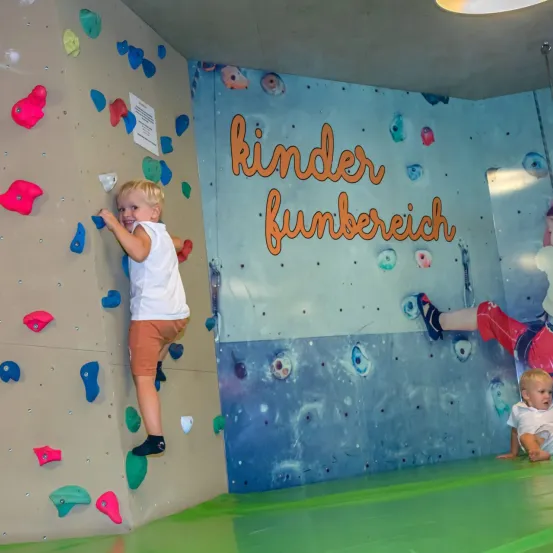 Ein junger Junge in Shorts und einem weißen Shirt klettert an einer Wand mit bunten Griffen. Die Wand ist mit 'Kinder Funbereich' beschriftet. Ein anderes Kind sitzt in der Nähe auf dem Boden.