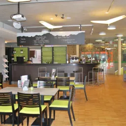 Ein modernes Indoor-Café mit Holzböden, einer schwarzen Theke, einer Tafel mit Speisekarte und verschiedenen Tischen und Stühlen. Flaschen und Weingläser sind auf der Theke ausgestellt.