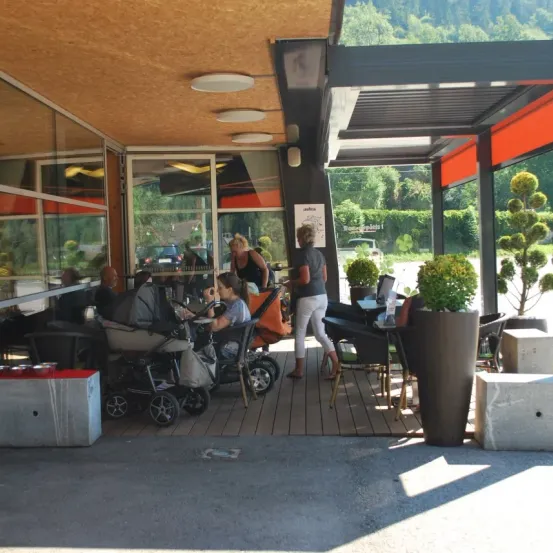 Ein Outdoor-Restaurant mit Familien, die an Tischen sitzen. Topfpflanzen und Markisen bieten Schatten. Bäume und ein Fahrzeug sind im Hintergrund zu sehen.