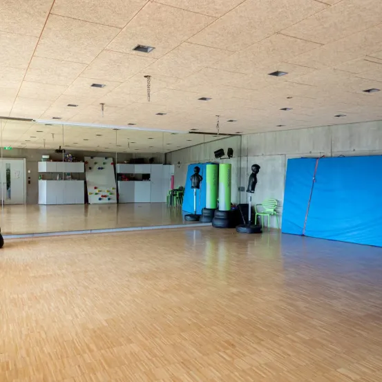 Ein leerer Fitnessraum mit einem großen Spiegel, Holzböden und verschiedenen Trainingsgeräten, einschließlich Reifen, grünen Zylindern und einem blauen Tarp.