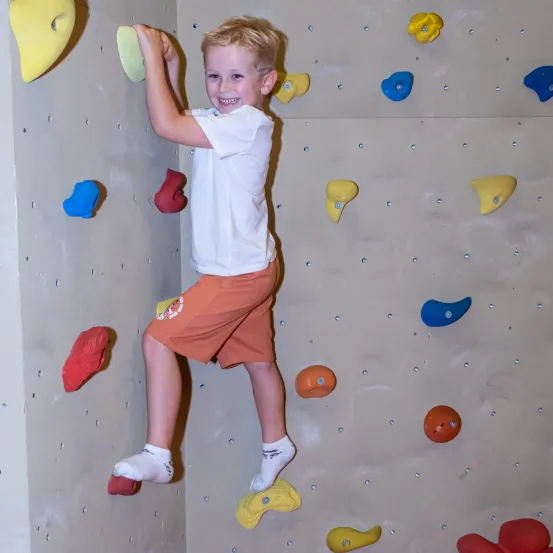 Ein junger Junge klettert an einer Wand mit bunten Klettergriffen. Er trägt ein weißes T-Shirt, orange Shorts und weiße Socken.