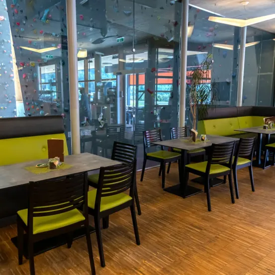 Ein Indoor-Restaurant mit Holzboden, schwarzen Stühlen und Tischen, mit einer Kletterwand mit farbigen Griffen.