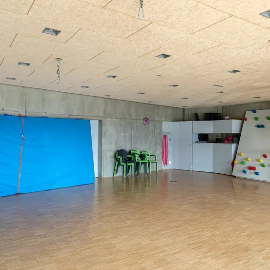 Ein leerer Raum mit einem blauen Hintergrund und einer Kletterwand mit bunten Blöcken. Es gibt grüne Stühle, die zur Seite gestapelt sind.