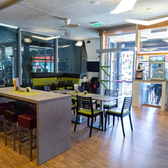 Ein Innenrestaurant mit modernen Möbeln und einem Holzboden. Tische und Stühle sind um eine zentrale Theke herum angeordnet. Glaswände bieten Ausblicke nach draußen.