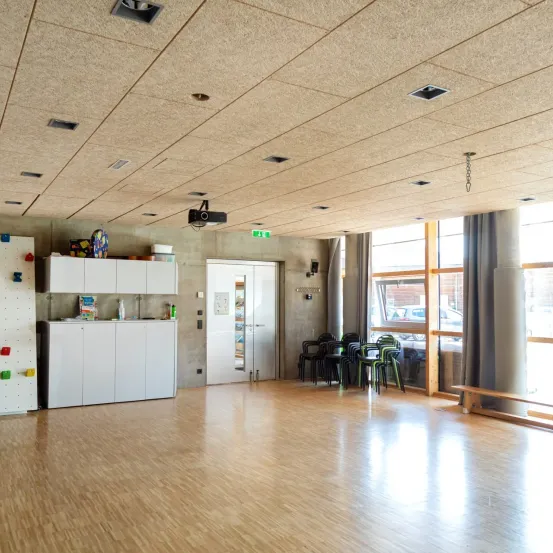 Ein leerer Raum mit Holzböden hat weiße Schränke und Regale auf der linken Seite. Ein Projektor ist an der Decke montiert. Stühle sind auf der rechten Seite angeordnet und ein Fenster ist an der Wand. Draußen ist ein Auto geparkt.