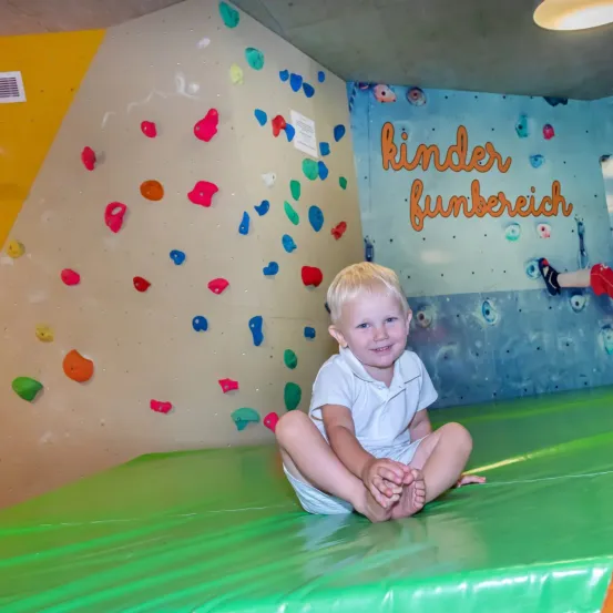 Ein junger Junge sitzt lächelnd auf einer grünen Matte in einem Indoor-Kletterpark. Die Wand hinter ihm ist mit bunten Klettergriffen gefüllt. Die Wand ist mit den Worten 'Kinder Funbereich' bemalt.