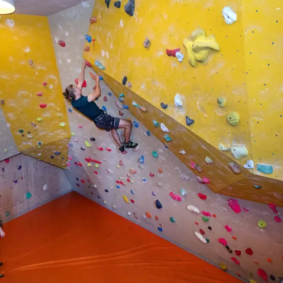 Eine Person klettert an einer gelben Kletterwand mit vielen farbigen Griffen und Haltepunkten, trägt ein grünes T-Shirt und Shorts. Die Wand ist geneigt und der Boden ist orange.
