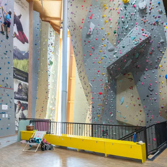 Ein Indoor-Kletterzentrum mit großen Wänden, die mit bunten Klettergriffen bedeckt sind. Zwei Kletterer erklimmen die Wände. Ein gelber Bank mit Taschen und einer blauen Flasche steht in der Nähe.