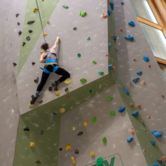 Eine Frau klettert an einer Kletterwand mit farbigen Griffen, trägt einen Gurt und Turnschuhe und greift einen gelben Griff.