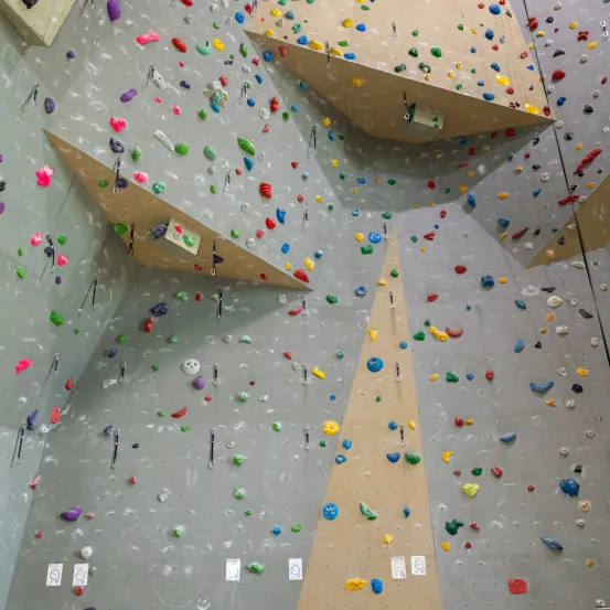 Eine Kletterwand, die mit mehrfarbigen Klettergriffen in verschiedenen Formen und Größen bedeckt ist, vor einem grauen Hintergrund mit dreieckigen Ausschnitten und weißen Griffaufklebern.