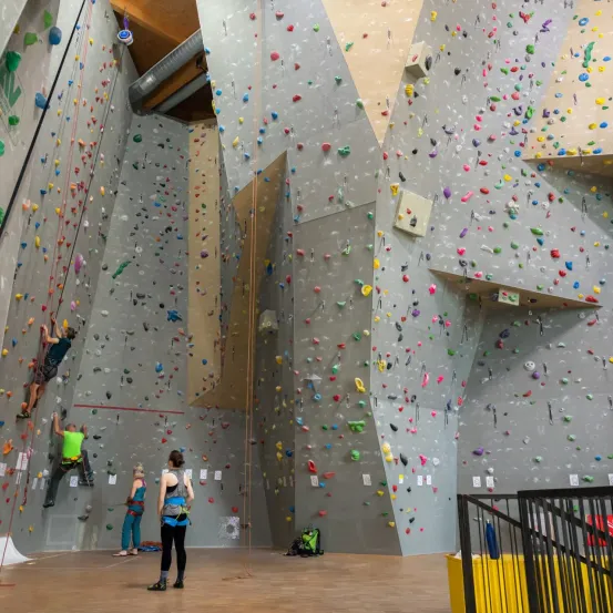 Indoor-Kletterhalle mit bunten Klettergriffen. Zwei Kletterer erklimmen verschiedene Wände. Drei Personen stehen am Boden, eine trägt ein grünes Shirt.