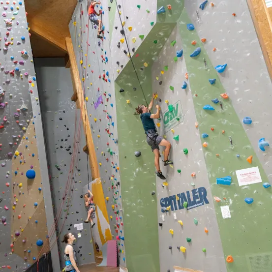 Mehrere Personen klettern drinnen. Eine Frau klettert an einer Wand, während eine andere Frau zusieht. Die Wände sind voller verschiedenfarbiger und geformter Steine.