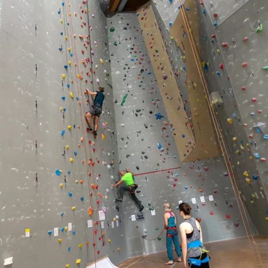 Zwei Personen klettern an einer Kletterwand mit vielen farbigen Griffen, während zwei andere unten zusehen.