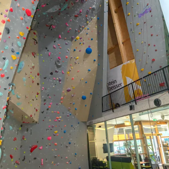 Ein Indoor-Kletterzentrum mit einer Wand voller bunter Klettergriffe und einem Balkon mit einem Banner, das 'stein beiss' liest.