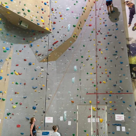 Eine Frau und ein Kind stehen vor einer Kletterwand, auf der eine Person klettert. Die Wand ist mit verschiedenen farbigen Klettergriffen bedeckt.