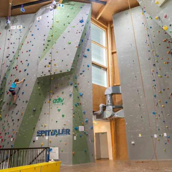 Ein Indoor-Kletterzentrum mit Kletterern, die bunte Wände erklimmen. Die Wände haben verschiedene Griffe in verschiedenen Farben und Formen. Ein Schild zeigt 'SPITALER'.