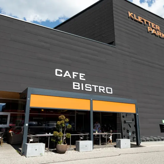 Der Eingang des Cafe Bistro, Teil des Kletter Parks, hat ein orangefarbenes Vordach, Sitzplätze im Freien und Topfpflanzen. Ein blauer Himmel mit Wolken ist oben sichtbar.