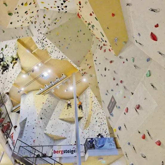 Eine Indoor-Kletterwand mit mehrfarbigen Klettergriffen und Wänden, mehreren Kletterern und einer Sicherheitsnetz. Es gibt Schilder und Lichter sowie eine Treppe, die in den oberen Stock führt.