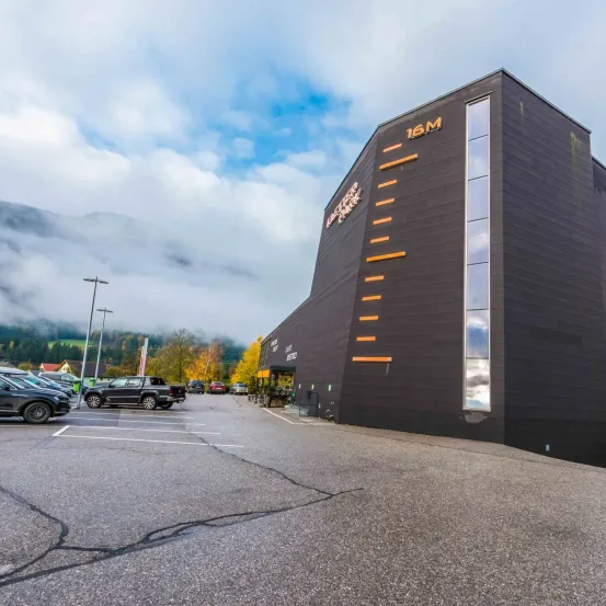 Ein schwarzes Gebäude mit gelber Schrift, gekennzeichnet mit 16M, steht prominent vor einem Bergpanorama. Fahrzeuge sind in einem Parkplatz vor dem Gebäude geparkt.