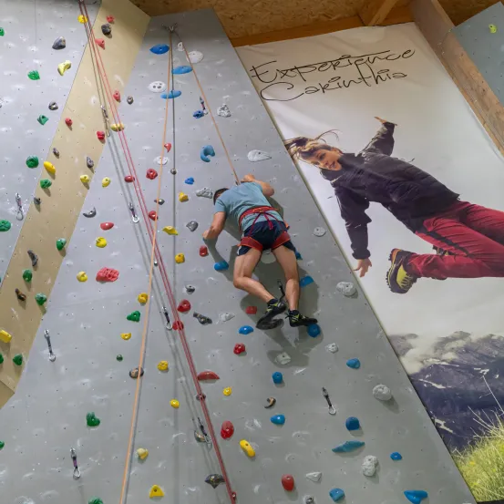 Eine Person klettert an einer Wand mit verschiedenen farbigen Griffen. Ein großes Poster hinter ihnen zeigt eine Person in ähnlicher Pose.