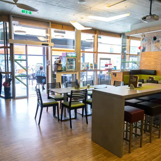 Ein Innenbereich zum Essen mit Holzböden und großen Glastüren. Tische und Stühle sind aufgestellt, und eine Person sitzt draußen auf einem Stuhl.