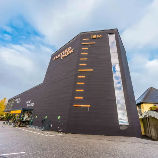 Das Äußere des Kletter Park Gebäudes ist schwarz mit orangefarbenen vertikalen Linien, mit den Texten 'Kletter Park' und '16M'. Eine Treppe befindet sich auf der rechten Seite.