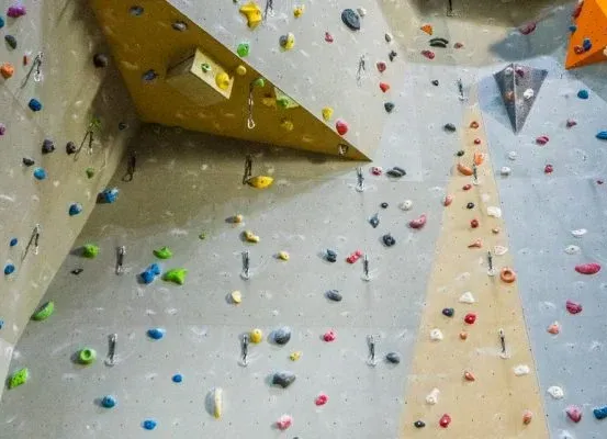 Eine Indoor-Kletterwand mit verschiedenfarbigen Griffen und Kletterern in der Ferne. Die Wand hat gelbe und orangefarbene Griffe, einige fehlen.