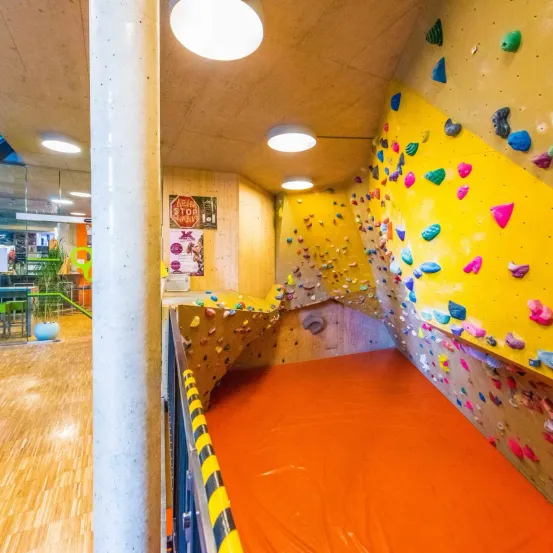 Ein Indoor-Klettergym mit einer gelben Wand, die mit Klettergriffen in verschiedenen Formen und Farben bedeckt ist. Der Boden ist orange und die Decke ist weiß mit Lichtern.
