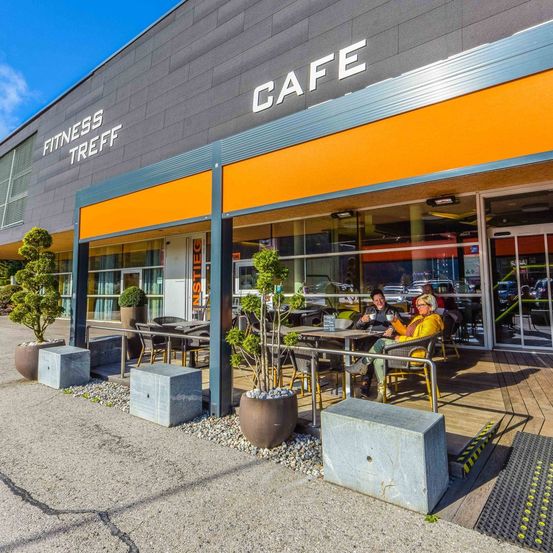 Ein Café im Freien, angeschlossen an ein Gebäude mit der Aufschrift 'Fitness Treff Cafe', mit Menschen, die an einem sonnigen Tag draußen sitzen.