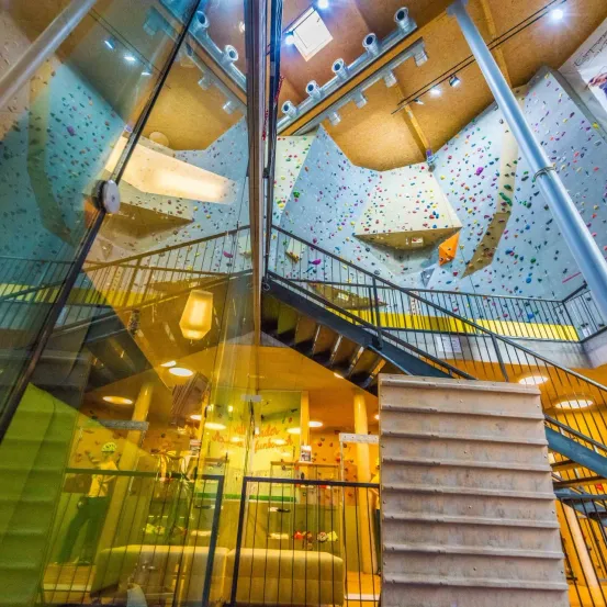 Indoor-Kletterhalle mit mehreren Kletterwänden, Treppen und Handläufen. Bunte Steine schmücken die Wände. Der Blick ist von einer Glaswand aus.