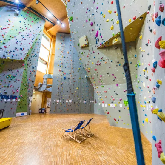 Ein leerer Indoor-Kletterpark mit Holzböden und Wänden, die mit bunten Klettergriffen bedeckt sind. Zwei blaue Stühle stehen auf dem Boden.