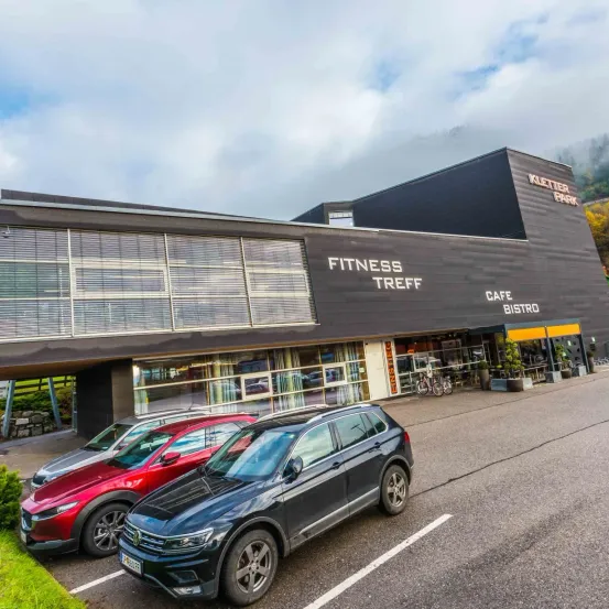 Ein Fitnesscenter mit Cafe und Bistro. Zwei Autos sind vor dem Gebäude geparkt. Das Gebäude hat Glasfenster und ein Schild, das Fitness Treff steht.