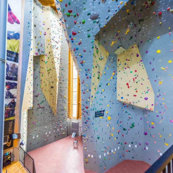 Eine Indoor-Kletterwand mit verschiedenen farbigen Klettergriffen und einem Poster an der linken Wand. Eine Bank steht in der Mitte des Raumes.