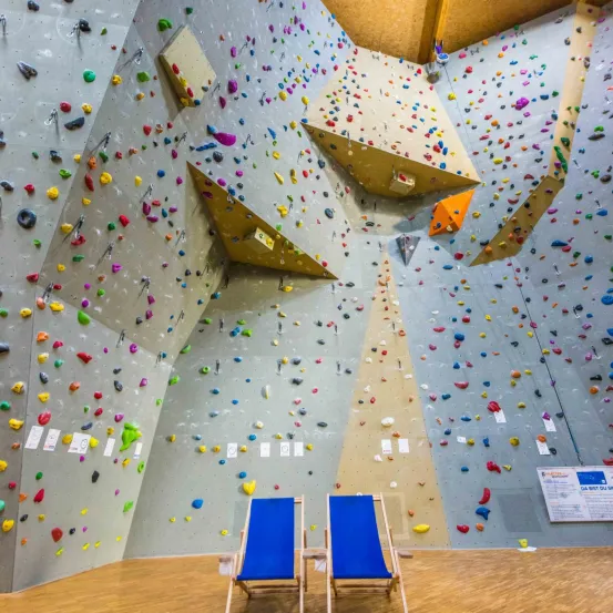 Eine Indoor-Kletterwand mit farbenfrohen Griffen und Haltegriffen, vor der zwei blaue Stühle stehen.