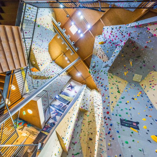 Eine Vogelperspektive einer mehrstöckigen Kletterhalle mit farbenfrohen Felsformationen, Klettergriffen und Sicherheitsschienen. Treppen und Metallhandläufe verbinden die Ebenen.