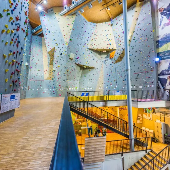 Ein großer Indoor-Kletterpark mit Holzböden und Kletterwänden, die mit farbenfrohen Griffen bedeckt sind. Treppen und Handläufe führen zu verschiedenen Ebenen. Werbung und Poster sind an den Wänden angebracht.