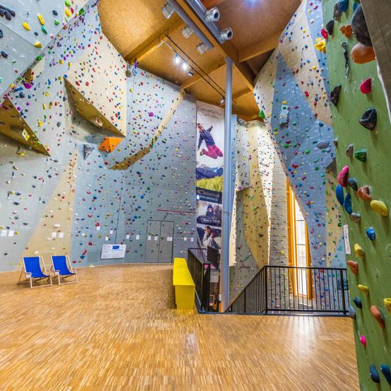 Ein Indoor-Kletterzentrum mit Wänden, die mit verschiedenen farbigen Klettergriffen bedeckt sind. Eine Person klettert an einer Wand. Zwei blaue Stühle stehen neben einer Wand und eine gelbe Bank neben den Stufen.