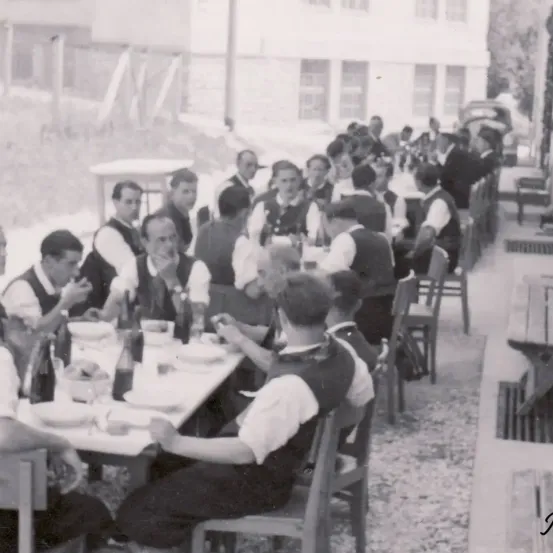 Ein Schwarz-Weiß-Foto einer Gruppe von Menschen, die um einen Tisch in einer Außenumgebung sitzen. Der Tisch ist mit Flaschen, Tellern und Schüsseln gefüllt.