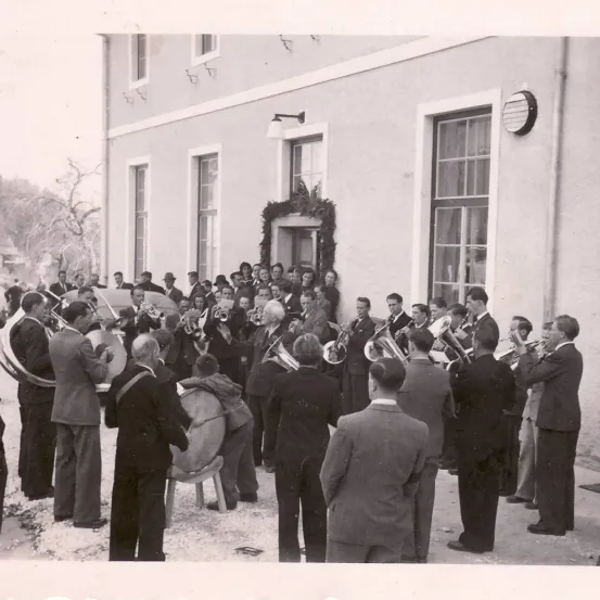 Ein Schwarz-Weiß-Foto einer Blaskapelle, die vor einem Gebäude auftritt, mit mehreren Zuschauern.