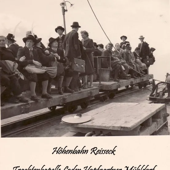 Ein Schwarz-Weiß-Foto von Menschen in altmodischer Kleidung, die auf einem Dampfzug sitzen. Die Gruppe lächelt und scheint die Fahrt zu genießen. Das Bild hat einen Wasserzeichen mit dem Text 'Hohenbahn Reisseck'.