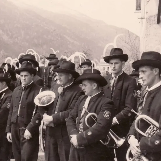 Eine Gruppe von Männern in formeller Kleidung, Hüten und Krawatten steht in einer Reihe, möglicherweise bei einer Parade, und hält Blechblasinstrumente. Die Umgebung scheint historisch zu sein.