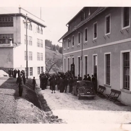 Ein altes Foto von Menschen, die vor einem Gebäude mit vielen Fenstern versammelt sind, einem geparkten Auto in der Nähe und Personen in formeller Kleidung.