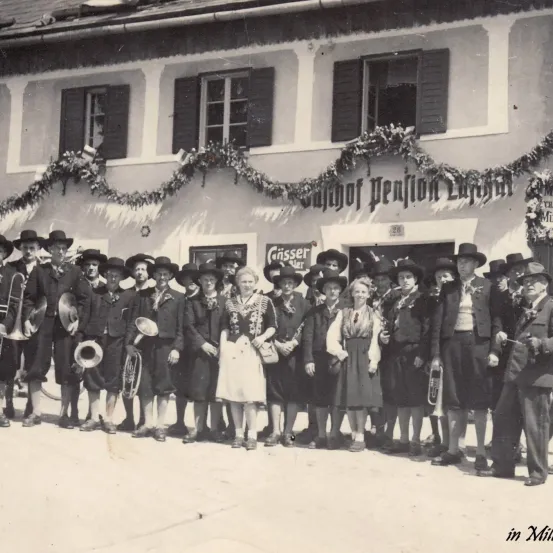 Ein Vintage-Foto einer Gruppe von Personen in traditioneller Kleidung, die Musikinstrumente halten und vor einem Gebäude mit dekorativen Kränzen posieren.
