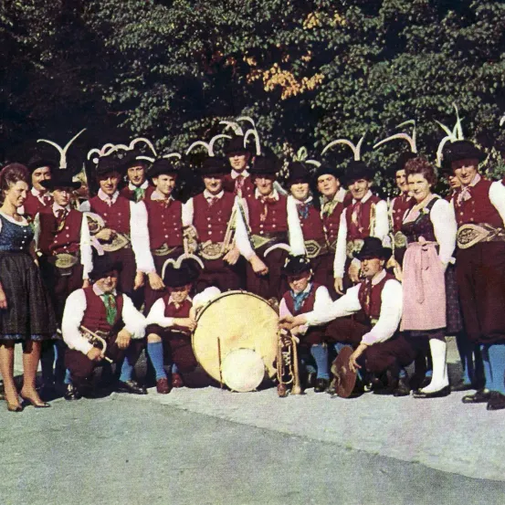 Ein altes Foto einer traditionellen Band, gekleidet in regionalen Kostümen, steht und sitzt in einem Halbkreis um eine Trommel. Sie halten Blechblasinstrumente und einige haben Hüte mit Hörnern. Bäume sind im Hintergrund.