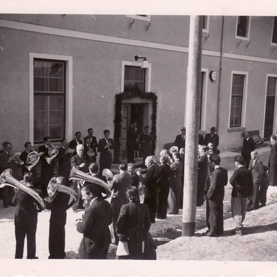 Ein Schwarz-Weiß-Foto einer Band, die vor einem Gebäude auftritt, mit mehreren Zuschauern, die zuschauen, und einigen, die in der Nähe eines Pfahls stehen.