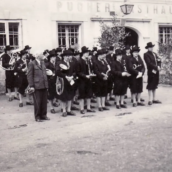 Eine Gruppe von Musikern in formeller Kleidung steht vor einem Gebäude und hält verschiedene Instrumente. Sie scheinen Teil einer Brassband zu sein.