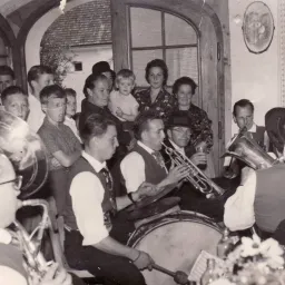 Ein Schwarz-Weiß-Foto zeigt eine Gruppe von Menschen, die drinnen versammelt sind und Musikinstrumente spielen. Die zentrale Figur hält eine Trompete, während andere Trommeln und Posaunen spielen. Eine Frau hält ein Kind und ein Mann trinkt aus einem Glas.